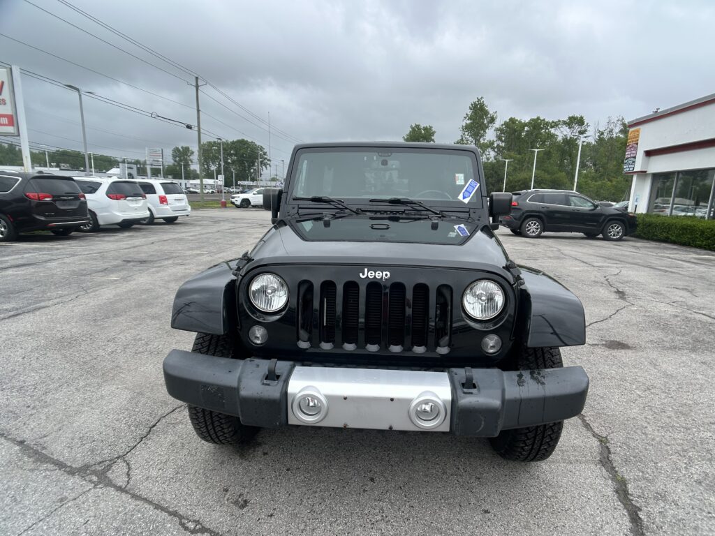 Front view of a black Jeep Wrangler parked in a car dealership lot with other vehicles and a white showroom building under a cloudy sky behind it