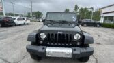 Front view of a black Jeep Wrangler parked in a car dealership lot with other vehicles and a white showroom building under a cloudy sky behind it