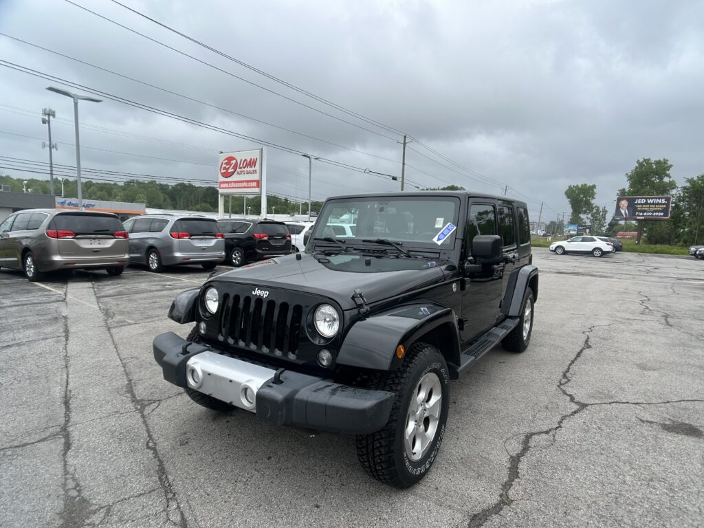 Front three-quarter view of a black Jeep Wrangler in a car dealership lot with other vans and an E-Z Loan Auto Sales sign nearby on a cloudy day.
