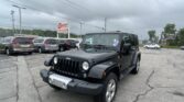 Front three-quarter view of a black Jeep Wrangler in a car dealership lot with other vans and an E-Z Loan Auto Sales sign nearby on a cloudy day.