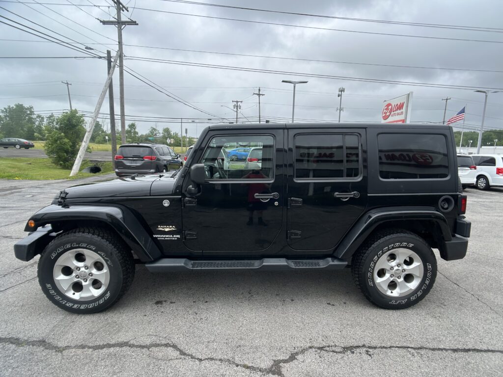 Black Jeep Wrangler Sahara in a parking lot with other cars and a cloudy sky; EZ Loan Auto Sales sign and American flag visible in the background