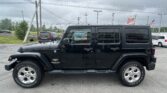 Black Jeep Wrangler Sahara in a parking lot with other cars and a cloudy sky; EZ Loan Auto Sales sign and American flag visible in the background