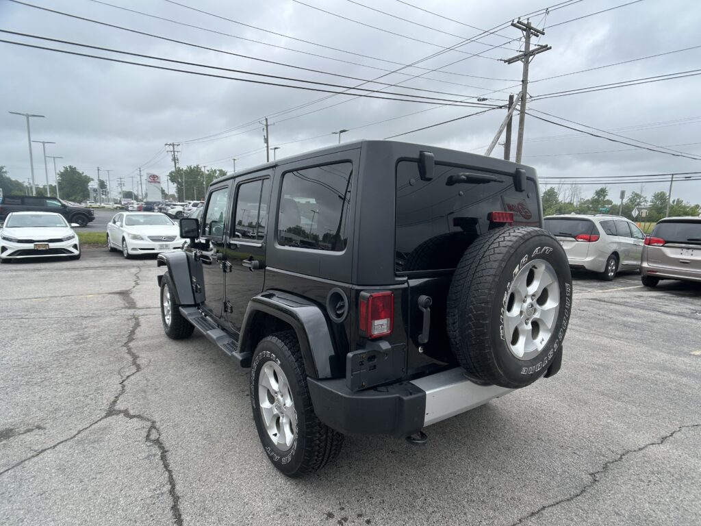 Rear three-quarter view of a black Jeep Wrangler with spare tire mounted on the tailgate in a crowded dealership lot under a cloudy sky