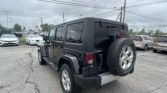 Rear three-quarter view of a black Jeep Wrangler with spare tire mounted on the tailgate in a crowded dealership lot under a cloudy sky
