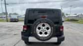 Rear view of a black Jeep with a mounted spare tire on the back in a parking lot, dealership buildings in the background and power lines overhead.