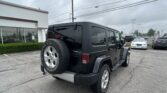Rear view of a black Jeep Wrangler with a full-size spare tire mounted on the back, parked in a lot beside a white building with red trim and overhead power lines."