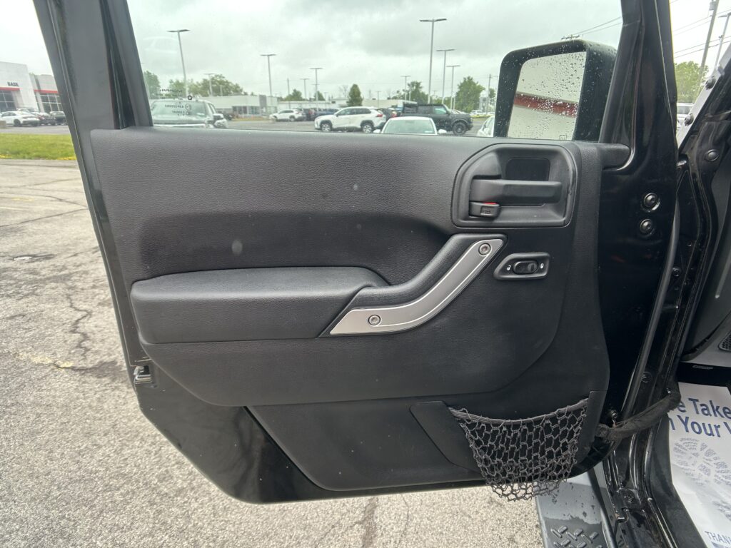 Close-up of a car door interior with a textured black panel, silver handle, and a net pocket; rainy parking lot visible outside.