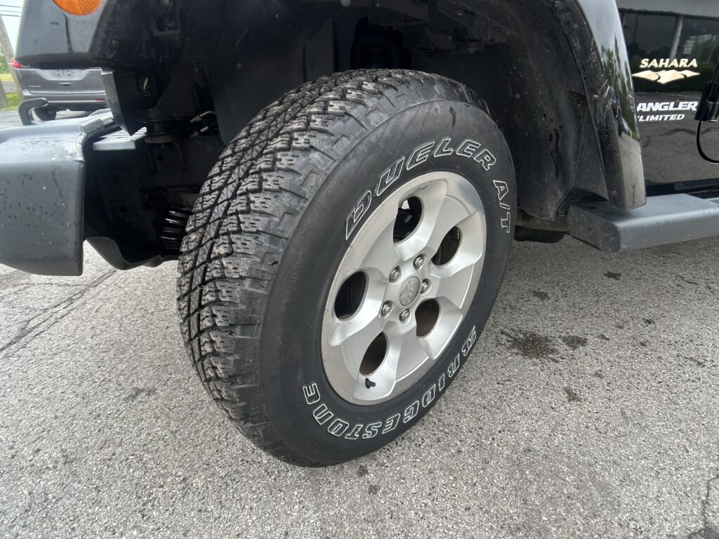 Close-up of a rugged all-terrain tire mounted on a silver wheel on a black Jeep Wrangler Sahara Unlimited, showing tread and fender.