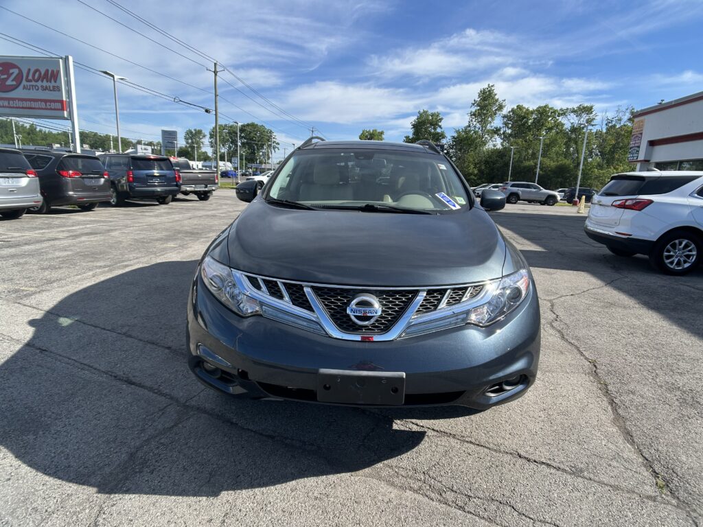 Front view of a gray Nissan SUV parked in a busy car lot with other vehicles and a dealership sign in the background under a blue sky.
