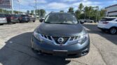 Front view of a gray Nissan SUV parked in a busy car lot with other vehicles and a dealership sign in the background under a blue sky.