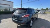 Rear view of a gray Nissan Murano SL AWD SUV in a sunny dealership lot with many cars and a white building on the left nearby menacing power lines above the scene