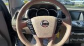 Driver's-seat view of a brown leather steering wheel with Nissan emblem; beige dashboard and center console, with a parking lot visible outside the windshield.