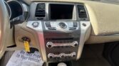 Front interior view of a car dashboard: center touchscreen, climate controls, gear shifter, and beige trim; outside a car lot visible through windshield.