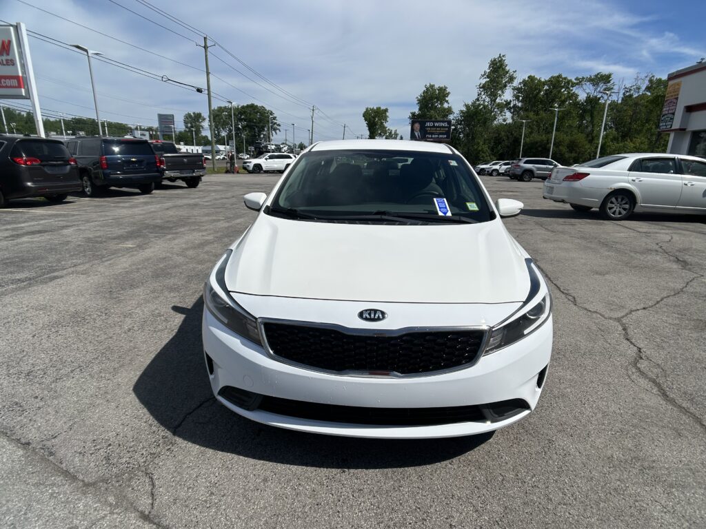 Front view of a white Kia sedan in a parking lot with other vehicles and a dealership lot in the background, daytime.