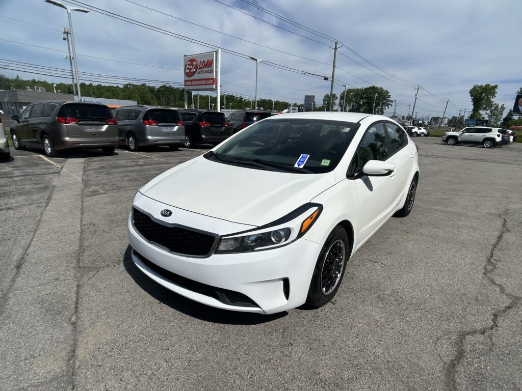 White Kia in the foreground at a used car lot with a row of minivans and an EZ Loan Auto Sales sign in the background.
