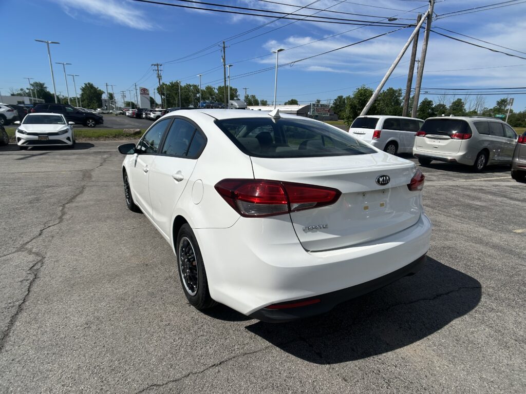 White Kia Forte in the foreground of a car dealership lot, with other vehicles and power lines overhead on a sunny day.
