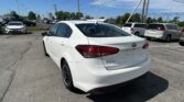 White Kia Forte in the foreground of a car dealership lot, with other vehicles and power lines overhead on a sunny day.