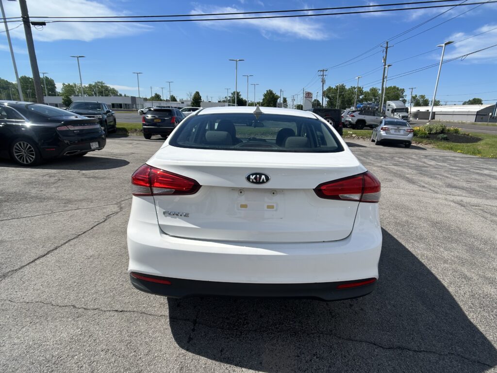 White Kia Forte parked in a car lot, viewed from behind, with other vehicles and a dealership in the background under a blue sky.