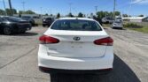 White Kia Forte parked in a car lot, viewed from behind, with other vehicles and a dealership in the background under a blue sky.