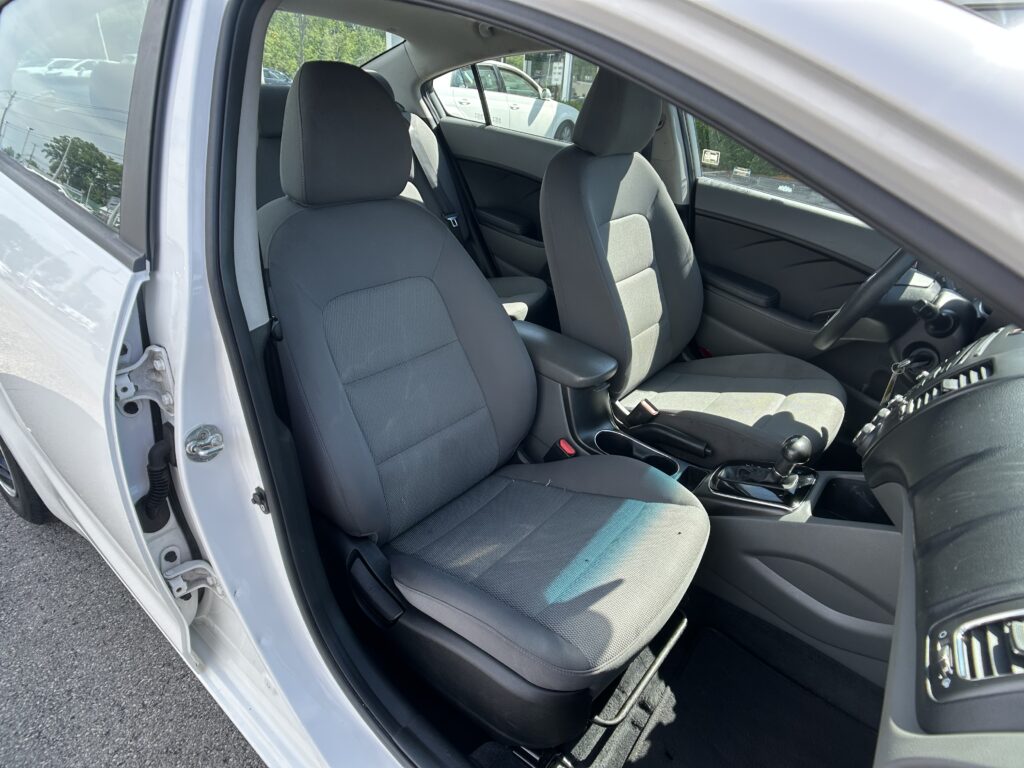 Interior of a gray-car cabin seen from open driver’s door: front seats, center console, and steering wheel visible in daytime.