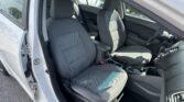 Interior of a gray-car cabin seen from open driver’s door: front seats, center console, and steering wheel visible in daytime.