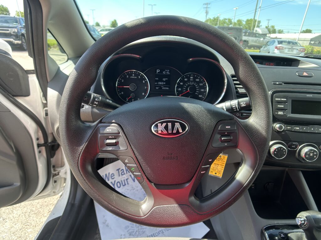 Inside a car cockpit from the driver's seat, steering wheel with Kia logo and dashboard gauges visible outside and center console to the right.