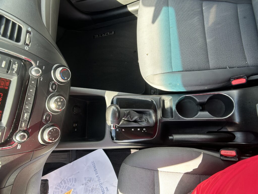 Interior of a car's front cabin showing the center console, gear shifter, climate controls, cup holders, and gray fabric seats.