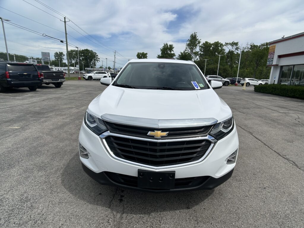Front view of a white Chevrolet SUV in a car dealership lot, with other vehicles and a building in the background.