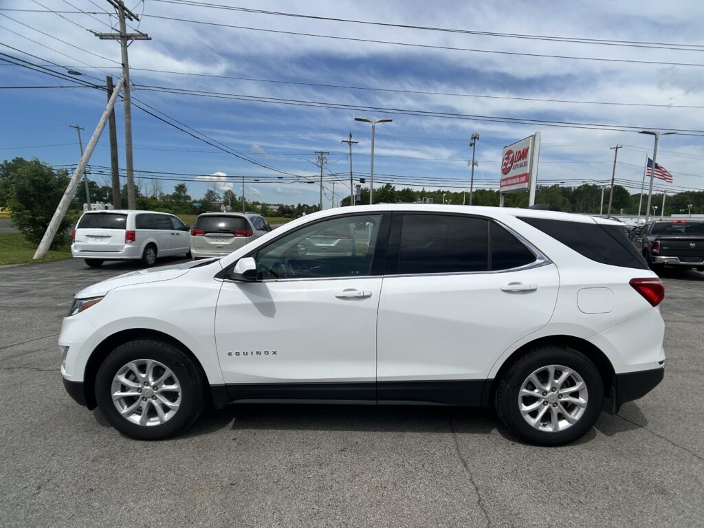 White Chevrolet Equinox SUV in a dealership lot with other cars, signs, and power lines against a blue sky with scattered clouds in the background