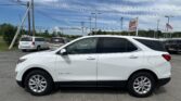 White Chevrolet Equinox SUV in a dealership lot with other cars, signs, and power lines against a blue sky with scattered clouds in the background