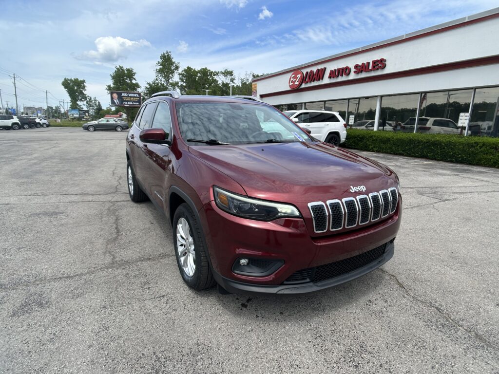 Red Jeep SUV in the foreground at a car dealership lot, with EZ Auto Sales building and other cars in the background under a blue sky.