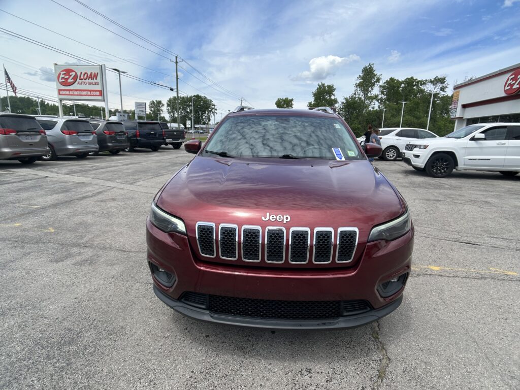 Close-up view of a red Jeep front grille and hood in a car dealer lot, with an E-Z Loan Auto Sales sign visible in the background.