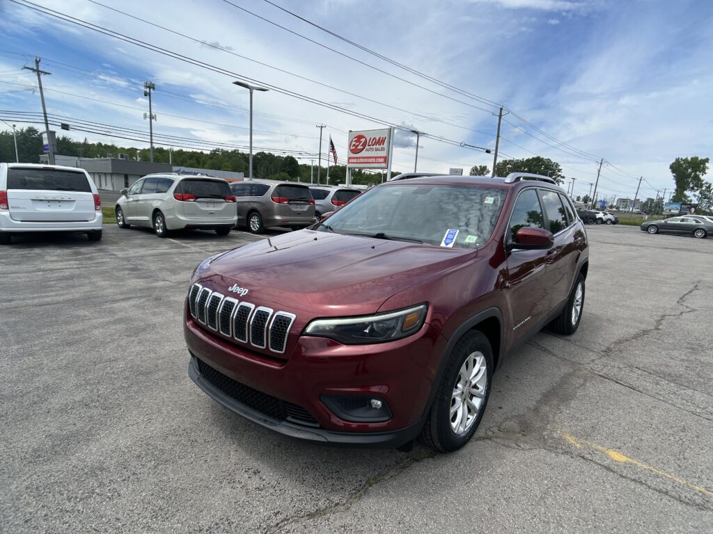 Maroon Jeep SUV in the dealership lot, with white minivans parked nearby and an EZ LOAN sign in the background under a blue sky.