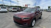Maroon Jeep SUV in the dealership lot, with white minivans parked nearby and an EZ LOAN sign in the background under a blue sky.