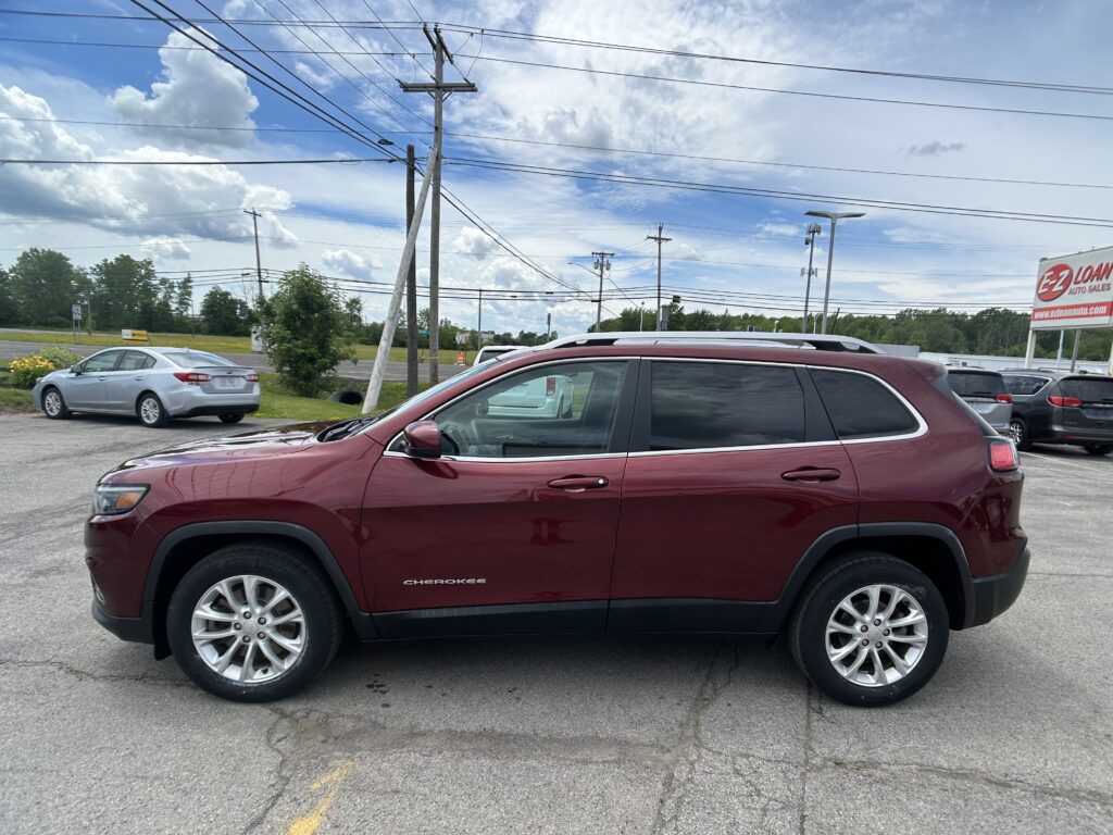 Red Jeep Cherokee SUV in a car dealership lot with other cars, power poles, and a blue cloudy sky above.