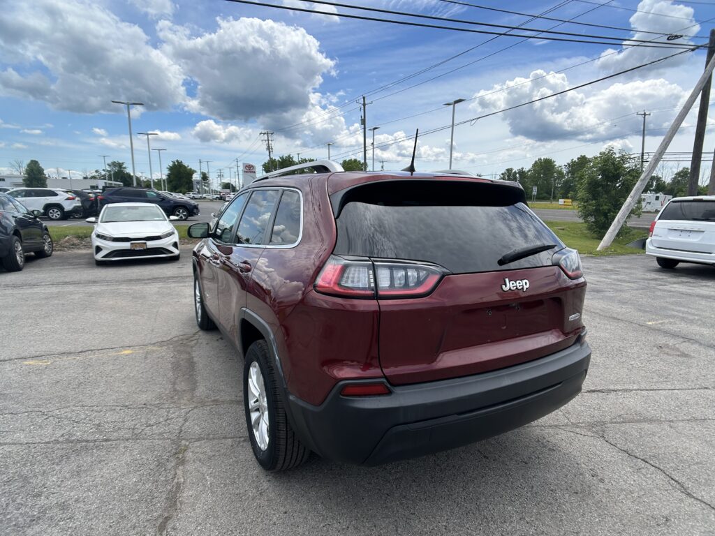 Rear view of a maroon Jeep SUV in a car dealership lot, with white and other vehicles and power lines against a cloudy blue sky behind.