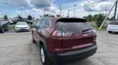 Rear view of a maroon Jeep SUV in a car dealership lot, with white and other vehicles and power lines against a cloudy blue sky behind.