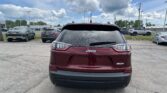 Rear view of a red Jeep Latitude SUV parked in a car lot, with other vehicles and a cloudy blue sky in the background