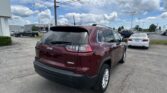 Rear view of a red Jeep Latitude SUV in a parking lot near a car dealership with other vehicles and a cloudy sky above.