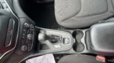 Front interior of a car showing the center console, gear shifter, cup holders, and fabric seats with papers on the floor.