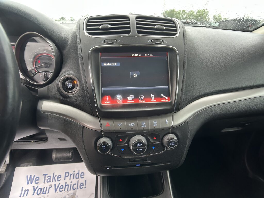 Center console car interior with touchscreen display, climate knobs, and start button; paper sign on floor reads 'We Take Pride In Your Vehicle!'