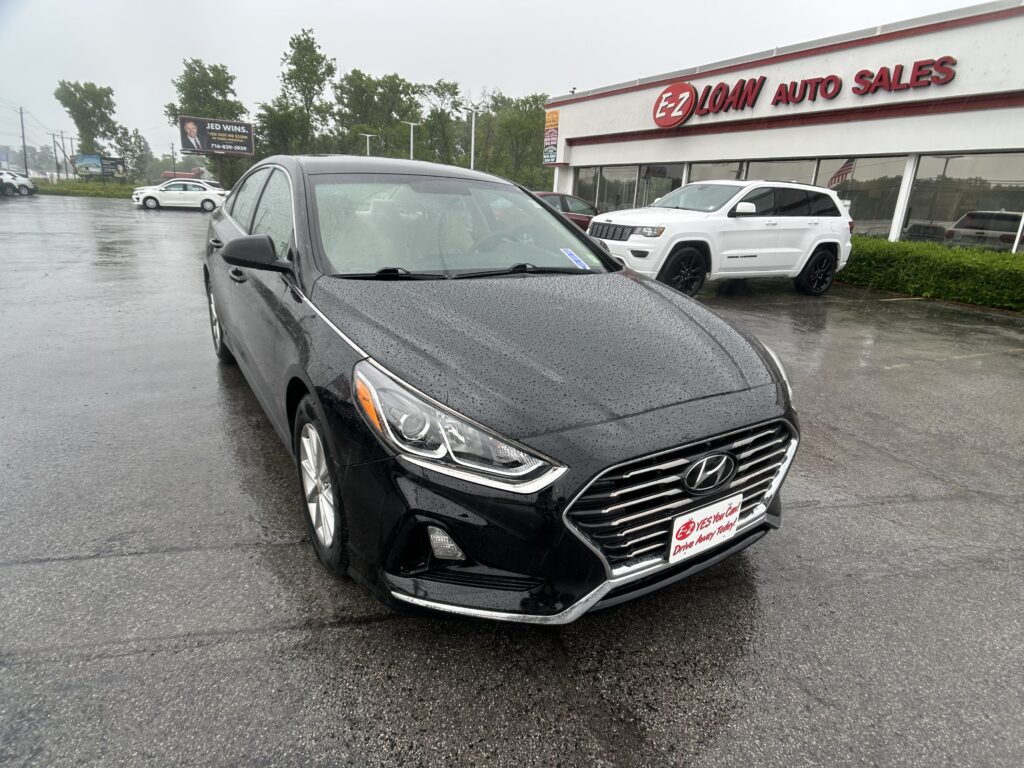 Black Hyundai sedan in a rain-soaked car lot in front of EZ Loan Auto Sales storefront with a white SUV nearby.
