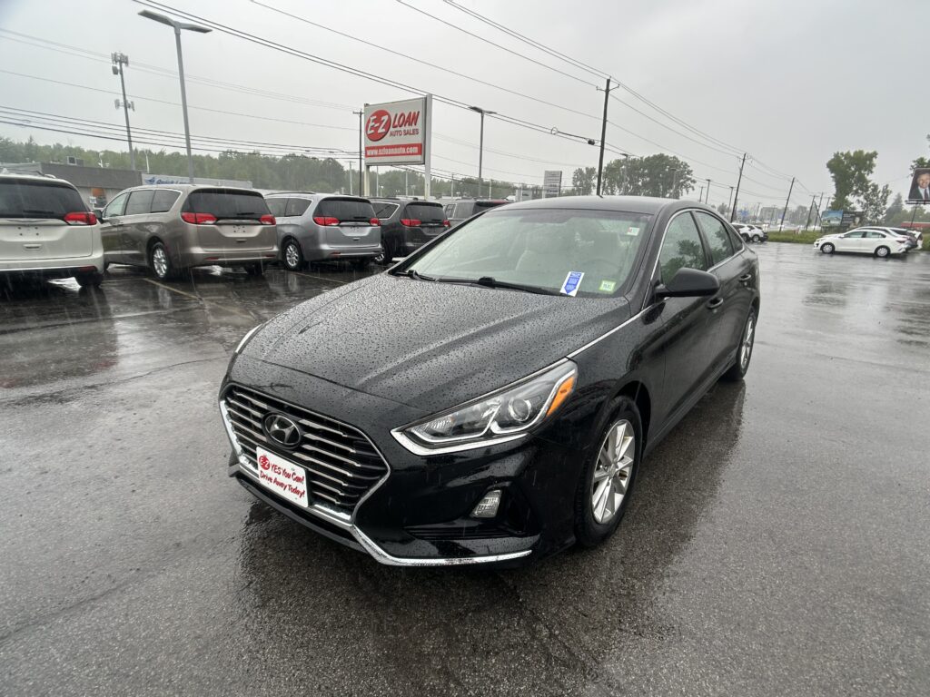 Black Hyundai sedan in the foreground at a rainy car dealership lot, with a row of minivans behind and an EZ LOAN Auto Sales sign in the background.