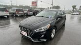 Black Hyundai sedan in the foreground at a rainy car dealership lot, with a row of minivans behind and an EZ LOAN Auto Sales sign in the background.