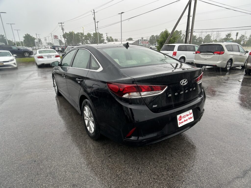 Rear view of a black Hyundai Sonata in a wet dealership lot with white and silver cars in the background during rain.