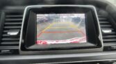 Car dashboard backup camera screen showing a parking lot with yellow guide lines and red boundary line; on-screen text reads 'Check surroundings for your safety'.