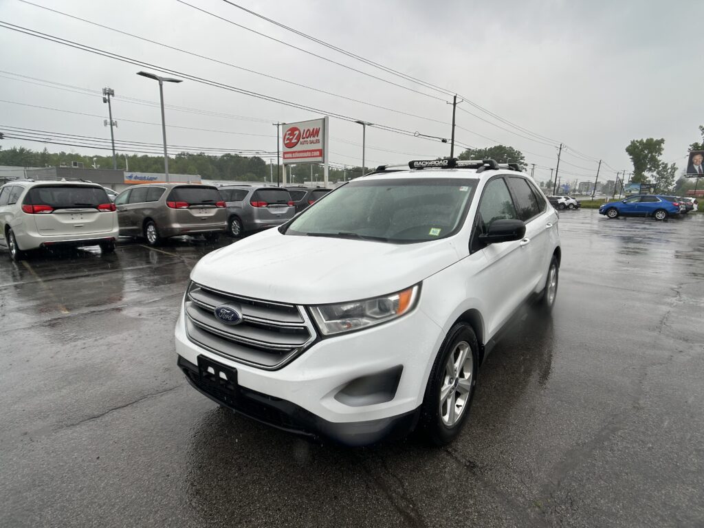 White Ford SUV in the foreground at a rainy used-car lot, with several minivans parked behind and power lines overhead near an EZ LOAN Auto Sales sign.