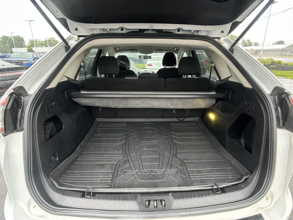 Open hatch shows a spacious SUV cargo area with a black rubber floor mat and rear seats upright, raindrops on windows in the background.