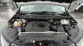 Open car hood revealing engine bay with coolant reservoir (left) and battery (right) in a rainy outdoor parking lot.