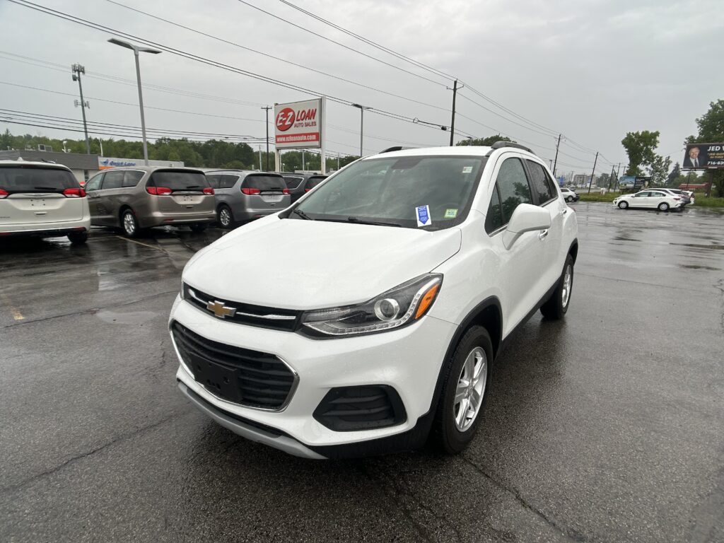 White Chevrolet SUV in a wet dealership lot with many minivans and an EZ LOAN sign in the distance.